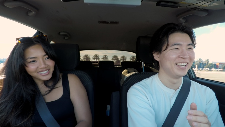 A student in the driver's seat and Yuji riding along, both laughing mid-lesson
